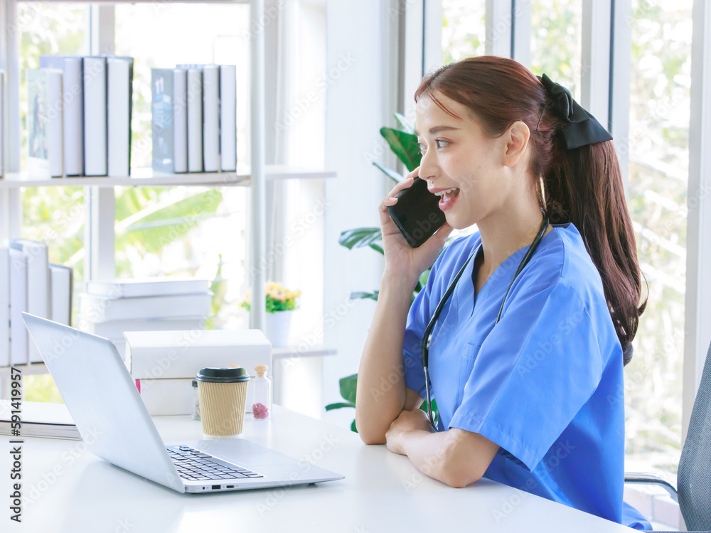 Asian young beautiful professional successful female doctor in blue uniform with stethoscope sitting smiling taking coffee break on call with smartphone in hospital office browsing laptop computer