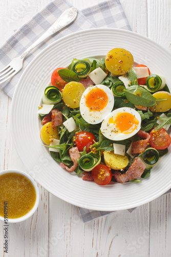 Peasant salad prepared with leafy vegetables, ham, cheese, tomatoes, potatoes and soft-boiled eggs closeup on the table. Vertical top view from above