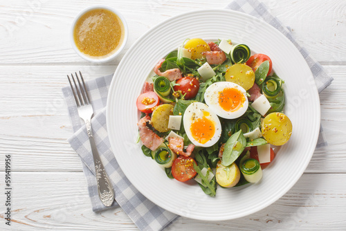 Farm salad with potatoes, tomatoes, bacon, arugula, zucchini, cheese and soft-boiled eggs seasoned with vinaigrette sauce close-up on a plate on the table. Horizontal top view from above photo