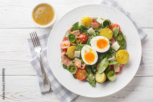 Fotografija Farm salad with potatoes, tomatoes, bacon, arugula, zucchini, cheese and soft-boiled eggs seasoned with vinaigrette sauce close-up on a plate on the table