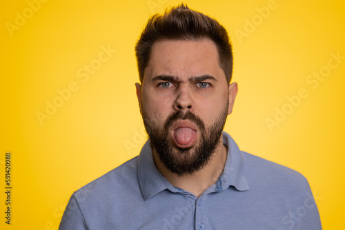 Young caucasian man showing tongue making childish faces at camera, fooling around, joking, aping with silly face, teasing, bullying abuse. Handsome guy isolated on studio yellow background indoors