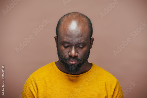 Portrait of African American man in yellow shirt looking down with sad expression standing on brown background
