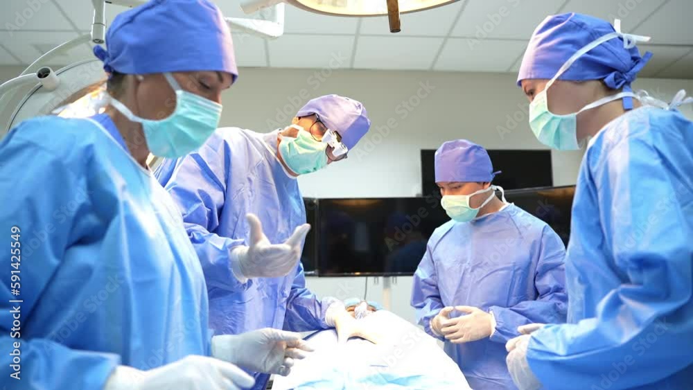 Zoom-out view of a surgical team in blue surgical uniforms and face masks, male surgeon asking for surgical instrument from nurse while surgery patient through a surgical drape in the operating room. Zoom-out view of a surgical team in blue surgical uniforms and face masks, male surgeon asking for surgical instrument from nurse while surgery patient through a surgical drape in the operating room.