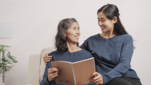 Reading a book, Mother's day concept, young adult female daughter congratulate excited asian elderly mother at sofa with birthday anniversary, two generations family photo, real people.