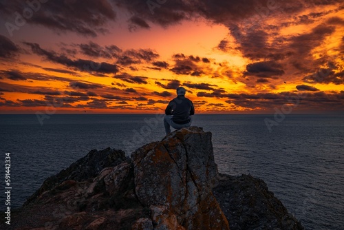 Rear of a male sitting on a pile of stones near seascape at sunset, dramatic sky background