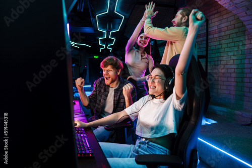 Group of friends celebrating victory of asian girl in video game while standing next to her in cybersport club