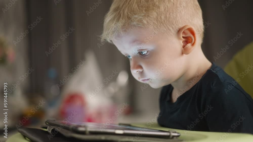 Blond toddler boy smiles while watches funny videos via tablet at home closeup. Cheerful little kid enjoys gadget. Internet technology of amusement Blond toddler boy smiles while watches funny videos via tablet at home closeup. Cheerful little kid enjoys gadget. Internet technology of amusement