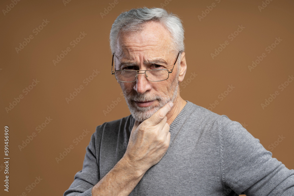 Closeup portrait of 60 years old, serious senior man wearing eyeglasses looking at camera isolated on brown background, holding hand near face. Vision concept Closeup portrait of 60 years old, serious senior man wearing eyeglasses looking at camera isolated on brown background, holding hand near face. Vision concept