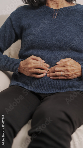 Asian mature woman holding her stomach, grimacing in pain. sitting on sofa.