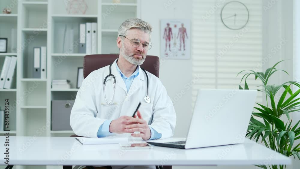 Mature doctor in white coat talking on video call using laptop while sitting at hospital clinic. Male medical worker physician has an online consultation with a patient sitting in a modern office Mature doctor in white coat talking on video call using laptop while sitting at hospital clinic. Male medical worker physician has an online consultation with a patient sitting in a modern office