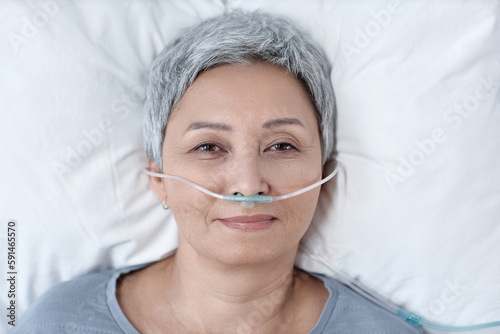 High angle view of senior woman in oxygen tube in her nose looking at camera while lying on bed in ward