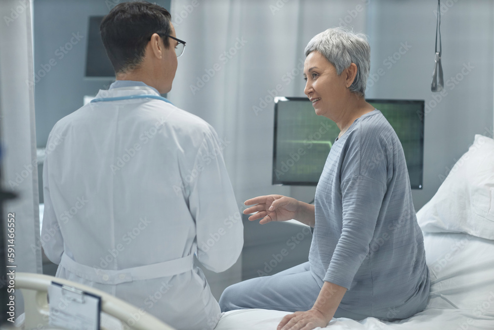Rear view of young doctor in white coat discussing health condition to senior patient while they sitting on bed in hospital ward
