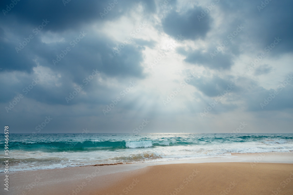 A gorgeous low-angle view of a sandy ocean beach with turquoise waves under a dramatic cloudy sky with sunlight through it.