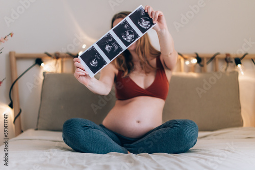 white, blonde pregnant woman sitting cross-legged on the bed shows pictures of the fetus taken with ultrasound photo