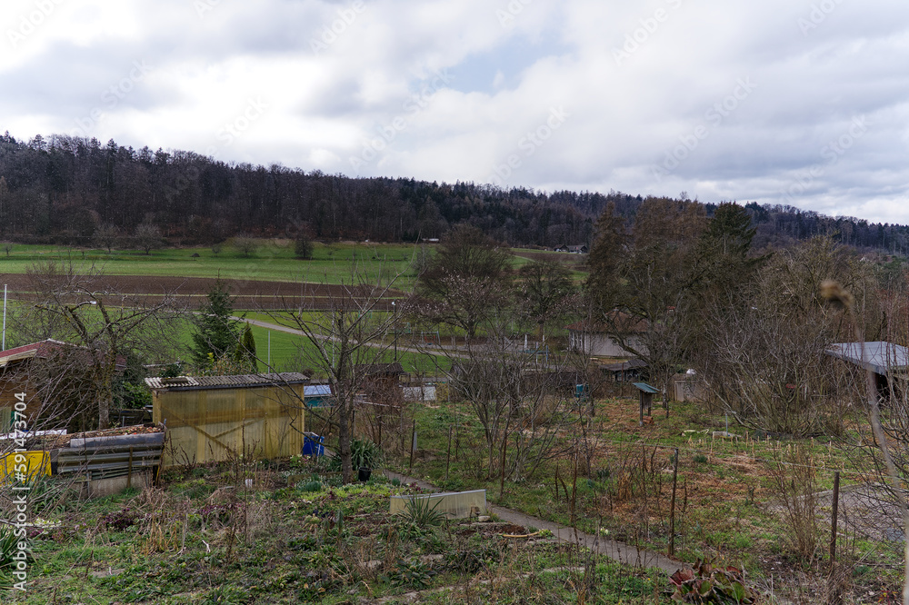 Fototapeta premium Scenic view over rural landscape and agriculture fields with garden plot in the foreground on a cloudy winter day. Photo taken February 26th, 2023, Zurich, Switzerland.
