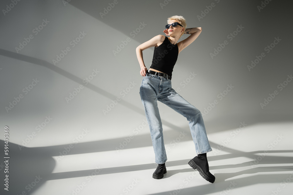 full length of tattooed model in black sunglasses and tank top posing on grey background.