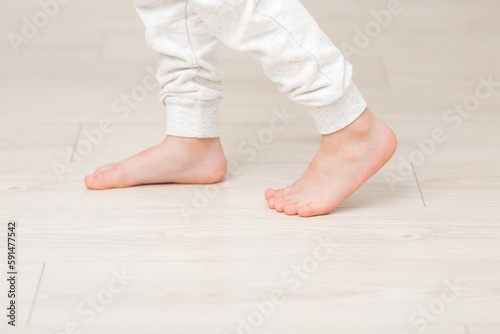 Little child legs in white trousers walking on light wooden floor background. Barefoot closeup. Side view.