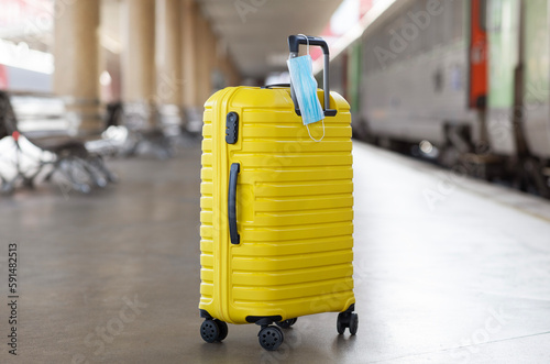 Yellow Suitcase With Hanged Protective Medical Face Mask Standing At Railway Station