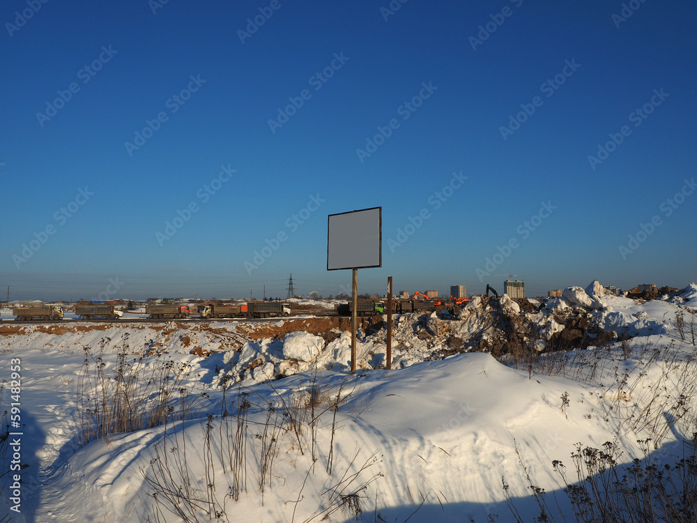 A sand pit covered with snow, trucks in the background, a stand with a white field for the inscription in the foreground. Construction machinery, road construction industry.