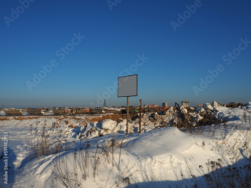 Obraz na płótnie A sand pit covered with snow, trucks in the background, a stand with a white field for the inscription in the foreground. Construction machinery, road construction industry. w salonie