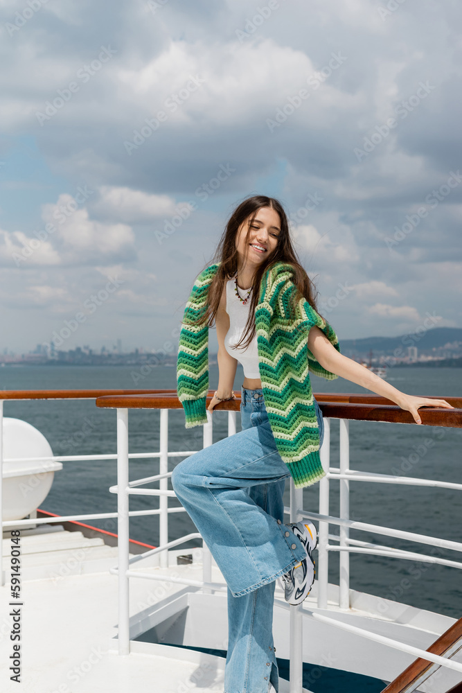 Positive young woman in knitted sweater and jeans standing near railing of yacht in Turkey.