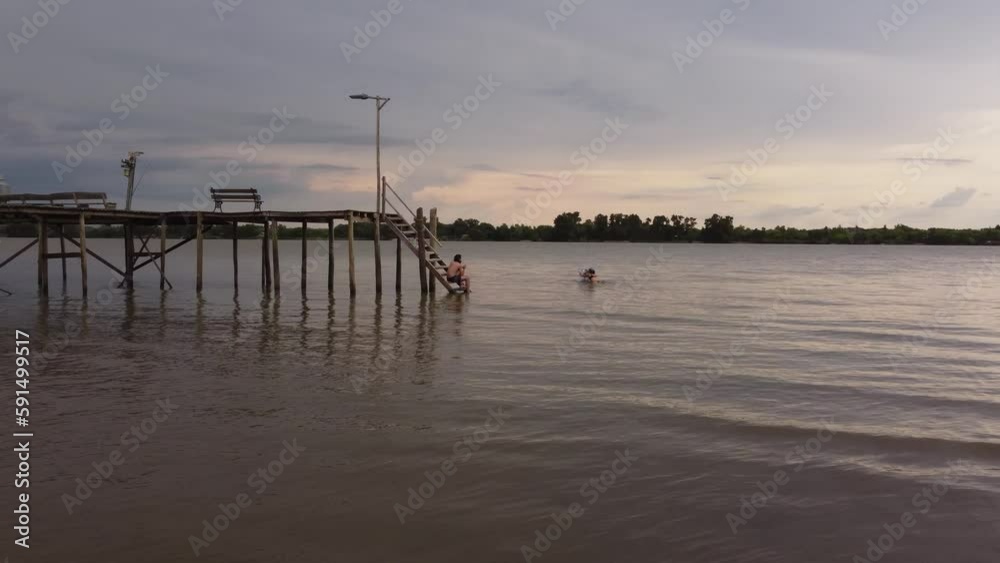 Couple of friends chilling in Parana river waters near wooden dock at sunset, Argentina. Aerial drone low Couple of friends chilling in Parana river waters near wooden dock at sunset, Argentina. Aerial drone low