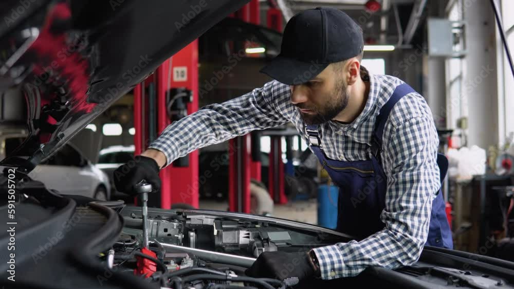 Auto mechanic examining broken car engine, standing near his car with open hood in a car repair shop