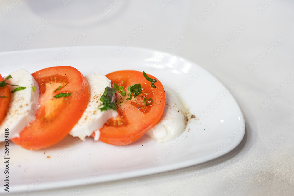 Caprese Salad with Mozzarella cheese, tomatoes, basil, olive oil on white plate at Italian diner, restaurant. Close up fresh Caprese is traditional Italian food from Italy. Traditional food concept. Caprese Salad with Mozzarella cheese, tomatoes, basil, olive oil on white plate at Italian diner, restaurant. Close up fresh Caprese is traditional Italian food from Italy. Traditional food concept.