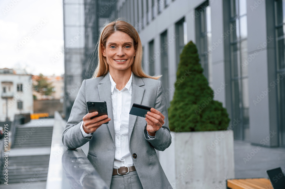 Holding credit card and smartphone. Woman in formal clothes is outdoors near the business building Holding credit card and smartphone. Woman in formal clothes is outdoors near the business building