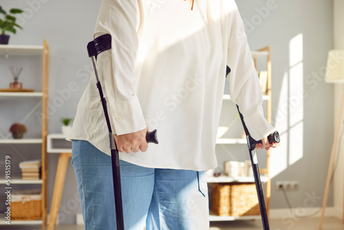 Injured overweight disabled woman using crutches to walk. Cropped shot of plump, plus size woman standing with axillary crutches in living room at home. Treatment and obesity problems