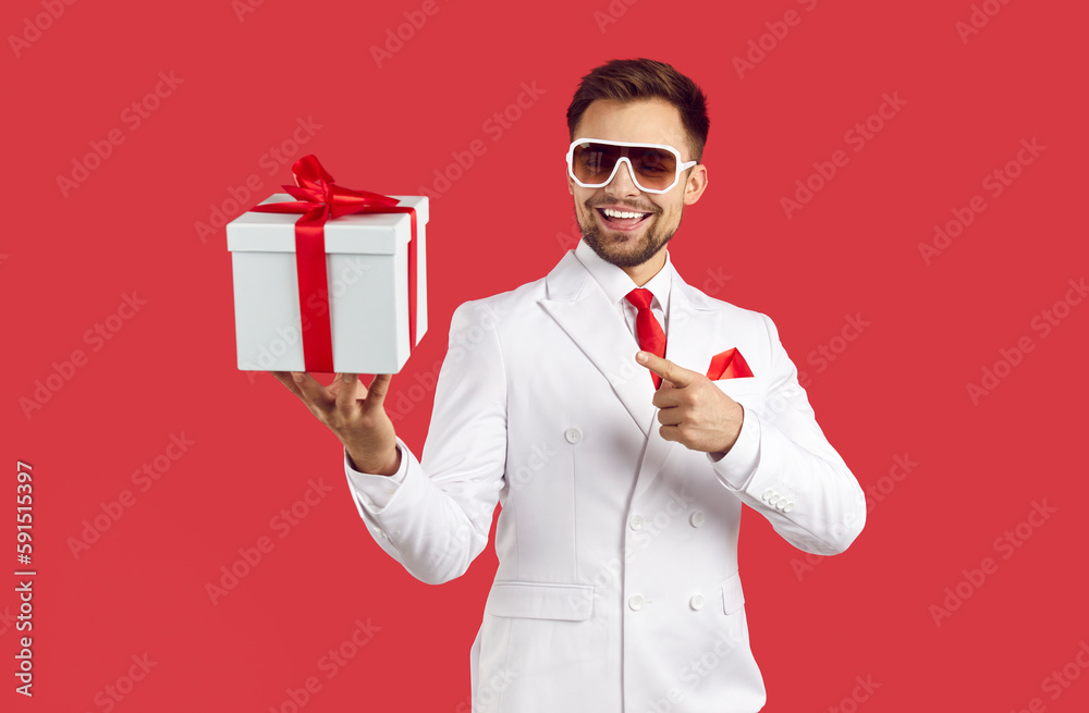 Cheerful stylish young man in suit points to gift box in his hands isolated on red background. Joyful man in white suit and sunglasses announces season of gifts and discounts. Advertising banner. Cheerful stylish young man in suit points to gift box in his hands isolated on red background. Joyful man in white suit and sunglasses announces season of gifts and discounts. Advertising banner.