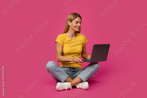 Cheerful middle aged european blonde female sitting on floor, typing on computer