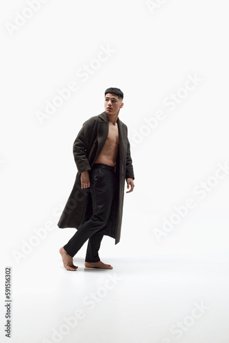 Full-length portrait of handsome, stylish, young, asian guy posing shirtless with trendy long coat against white studio background. Concept of fashion, body aesthetics, beauty, men's health, emotions