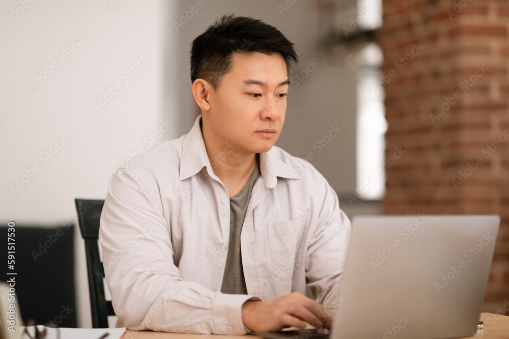 Remote job concept. Focused middle aged asian man sitting at workdesk at home, using laptop, working online Remote job concept. Focused middle aged asian man sitting at workdesk at home, using laptop, working online