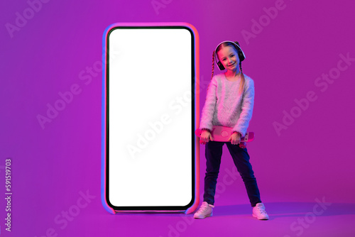 Sweet school-age girl with wireless headphones and skateboard next phone