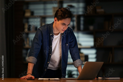 Positive young man manager standing by workdesk, looking at laptop