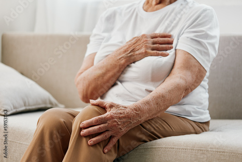 Elderly woman severe chest pain sitting on the sofa, health problems in old age, poor quality of life. Grandmother with gray hair holds her chest with her hands, women's health, breast cancer.