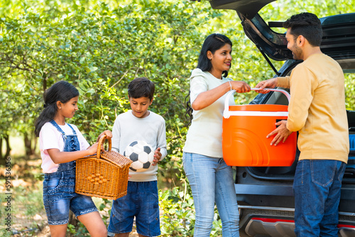 Happy Indian couple with kids taking out bags and picnic bucket from car boot - concept of summer holidays, togetherness and family vacation photo