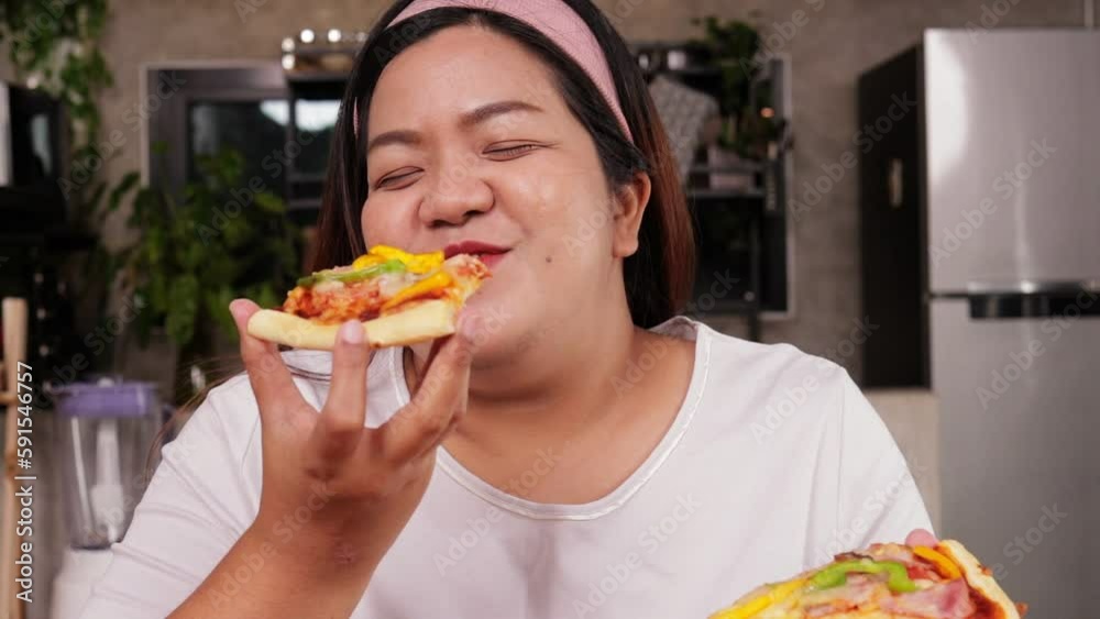 Fat Asian happy woman eating pizza, she holds two slices of pizza and eats deliciously in the kitchen at home. Fast food concept. weight loss Fat Asian happy woman eating pizza, she holds two slices of pizza and eats deliciously in the kitchen at home. Fast food concept. weight loss