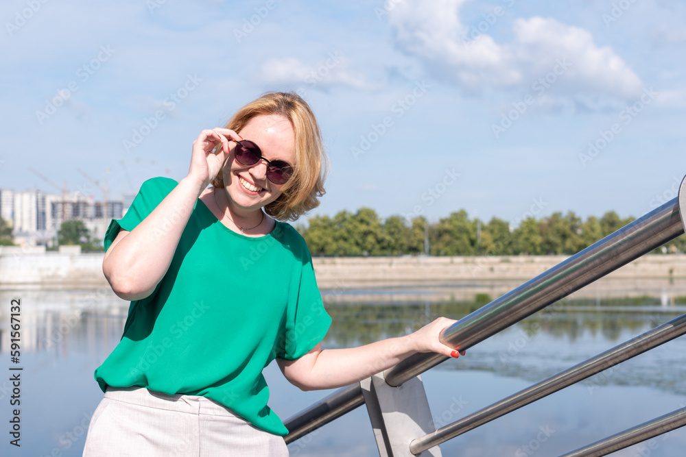 Portrait of a smiling happy girl in sunglasses, green blouse standing on the river bank.