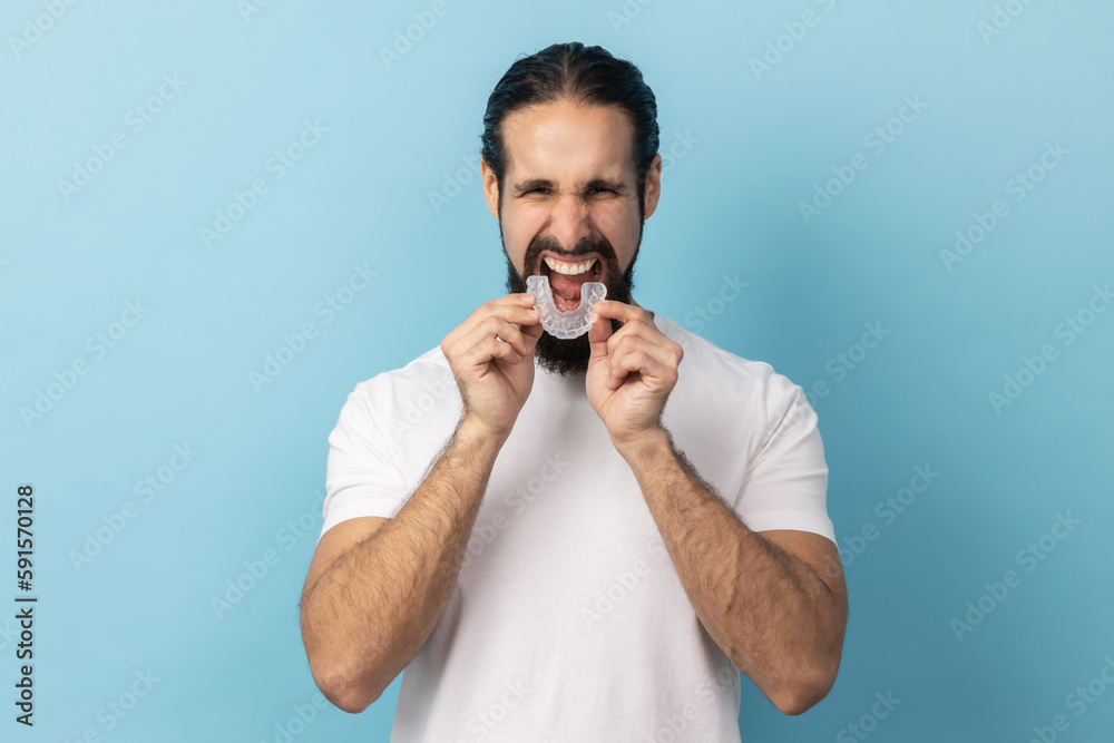 Portrait of man with beard wearing white T-shirt putting dental aligner retainer, dental clinic for beautiful teeth, treatment course concept. Indoor studio shot isolated on blue background. Portrait of man with beard wearing white T-shirt putting dental aligner retainer, dental clinic for beautiful teeth, treatment course concept. Indoor studio shot isolated on blue background.