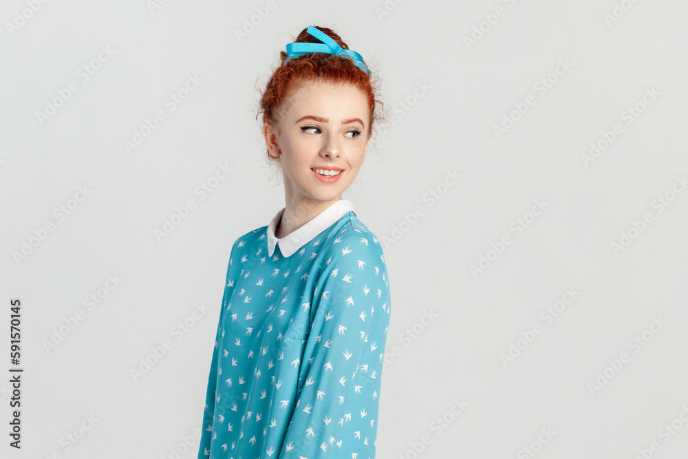 Side view portrait of charming red haired woman with bun hairstyle, standing looking away with pleasant smile, expressing happiness, wearing blue dress. Indoor studio shot isolated on gray background. Side view portrait of charming red haired woman with bun hairstyle, standing looking away with pleasant smile, expressing happiness, wearing blue dress. Indoor studio shot isolated on gray background.