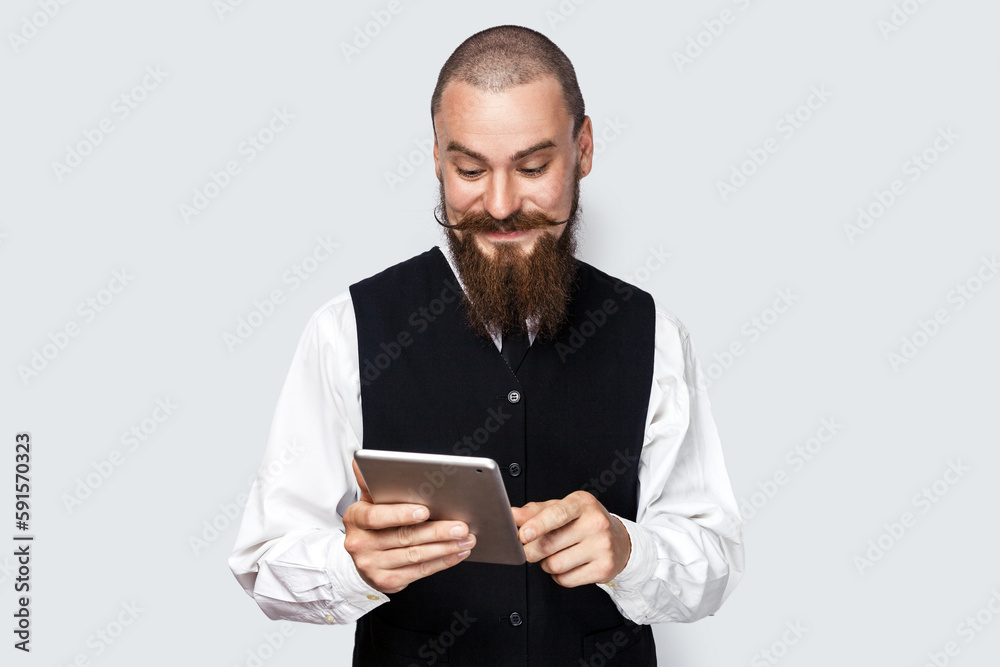 Portrait of smiling optimistic handsome man with beard and mustache standing using tablet, chatting with friends, reading message in social network. Indoor studio shot isolated on gray background. Portrait of smiling optimistic handsome man with beard and mustache standing using tablet, chatting with friends, reading message in social network. Indoor studio shot isolated on gray background.