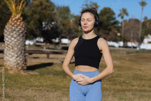 Concentrated young woman wearing headphones doing yoga meditation with online tutorial outdoors at park with palm trees. Listening music, harmony, calming, spirituality concept. World health day