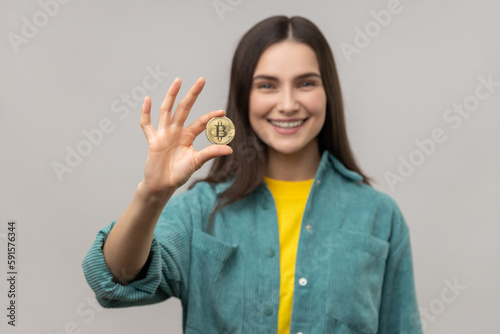 Portrait of smiling optimistic woman with dark hair holding gold bitcoin and looking at camera with toothy smile, wearing casual style jacket. Indoor studio shot isolated on gray background.