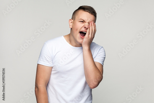 Portrait of sleepy overworked handsome teenager boy wearing T-shirt having not enough sleep, yawning, covering half of face with palm. Indoor studio shot isolated on gray background.