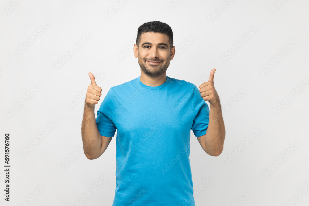 Portrait of optimistic cheerful smiling unshaven man wearing blue T- shirt standing showing thumb up, recommend something, good feedback. Indoor studio shot isolated on gray background. Portrait of optimistic cheerful smiling unshaven man wearing blue T- shirt standing showing thumb up, recommend something, good feedback. Indoor studio shot isolated on gray background.