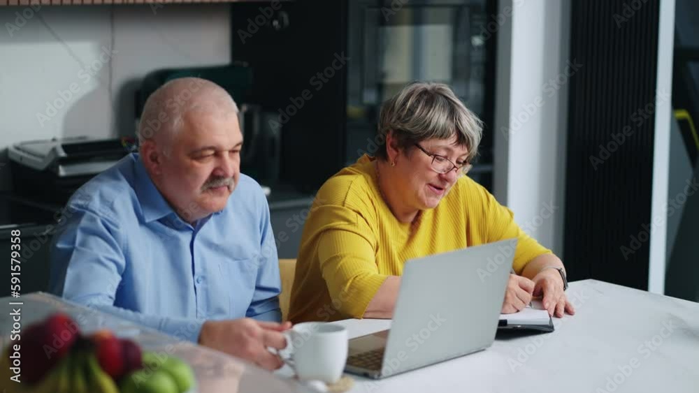 Pair Of Aged Spouses Checking Utility Bills Online On Display Of Laptop, Sitting Together In Kitchen Pair Of Aged Spouses Checking Utility Bills Online On Display Of Laptop, Sitting Together In Kitchen