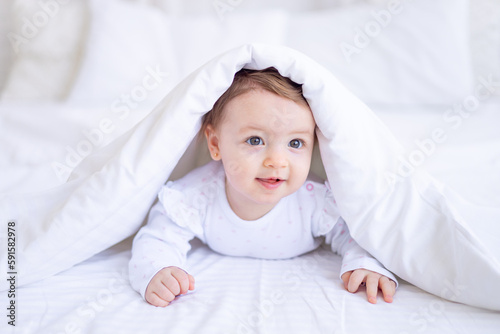 funny happy baby girl looks out from under the blanket on the bed in white clothes and smiles, a small child on a cotton bed at home woke up in the morning after sleeping