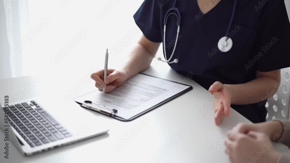 Doctor and patient sitting and discussing test results at the white desk near flair window in clinic. Unknown female physician use clipboard for filling up medical record form. Medicine concept Doctor and patient sitting and discussing test results at the white desk near flair window in clinic. Unknown female physician use clipboard for filling up medical record form. Medicine concept